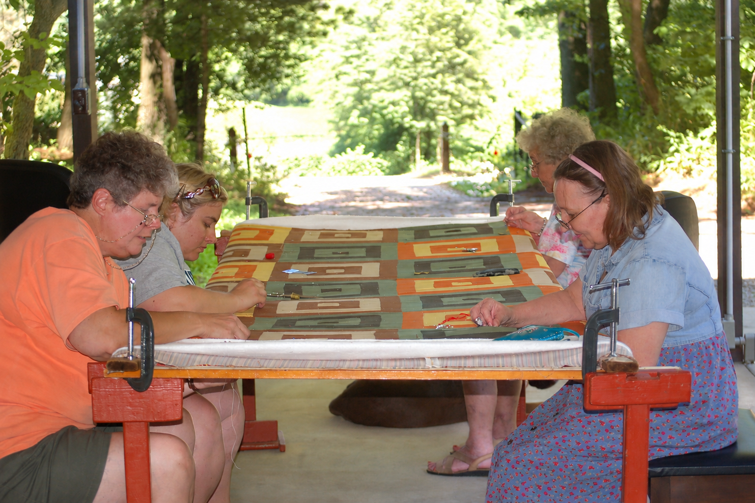 Four women gathered around a hand-quilting frame, quilting together outdoors